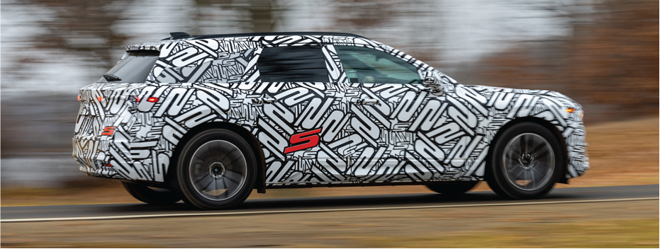 ¾ rear side view of ZDX with a black and white "S" monogrammed paint job driving down lakeside highway. The trees in the background are blurry.