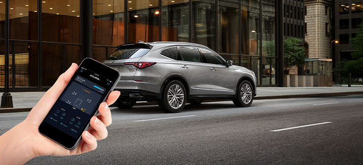 In the foreground, a hand is holding a smartphone displaying the AcuraLink™ app. In the background, a grey MDX is parked on a city street. / À l’avant-plan, une main tient un téléphone intelligent affichant l’application AcuraLinkMC. En arrière-plan, un MDX gris est stationné sur une rue urbaine.