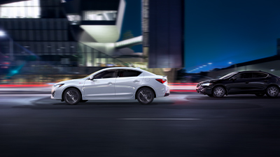 Side view of a white Acura at night with headlights on. / Vue latérale d’une Acura blanche la nuit, avec phares allumés. 