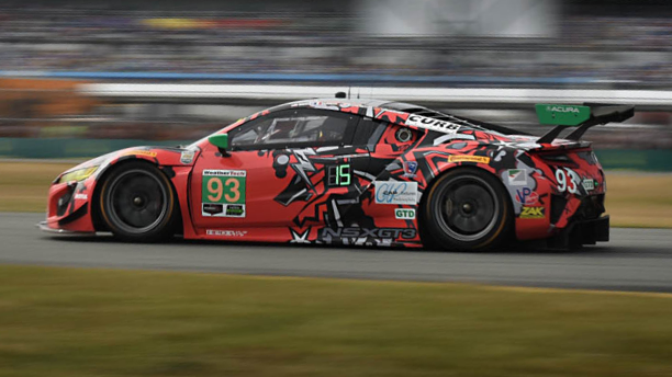 Side view of a red Acura NSX GT3  wrapped in sponsorship logos, driving at high speed on a racetrack, the stad ium blurred in the background.	Vue latérale d’une Acura NSX GT3 rouge  recouverte de logos de commanditaires roulant à haute vitesse sur un circuit de course avec des estrades en arrière-plan.