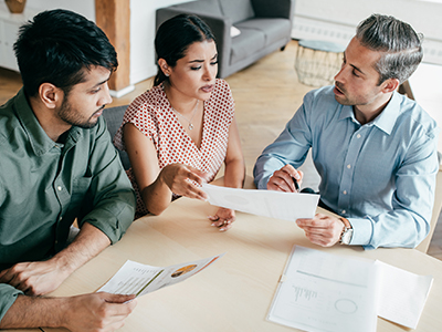Three people sit around a table discussing paperwork. / Trois personnes sont assises autour d'une table pour discuter au sujet de documents.